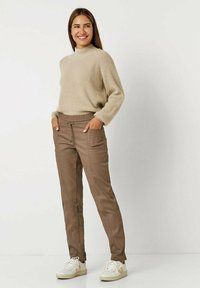 Beige Strickpullover mit hohem Kragen, kombiniert mit braunen schmal zulaufenden Hosen mit Taschen und verstellbarem Bund, abgerundet mit weißen Sneakers.