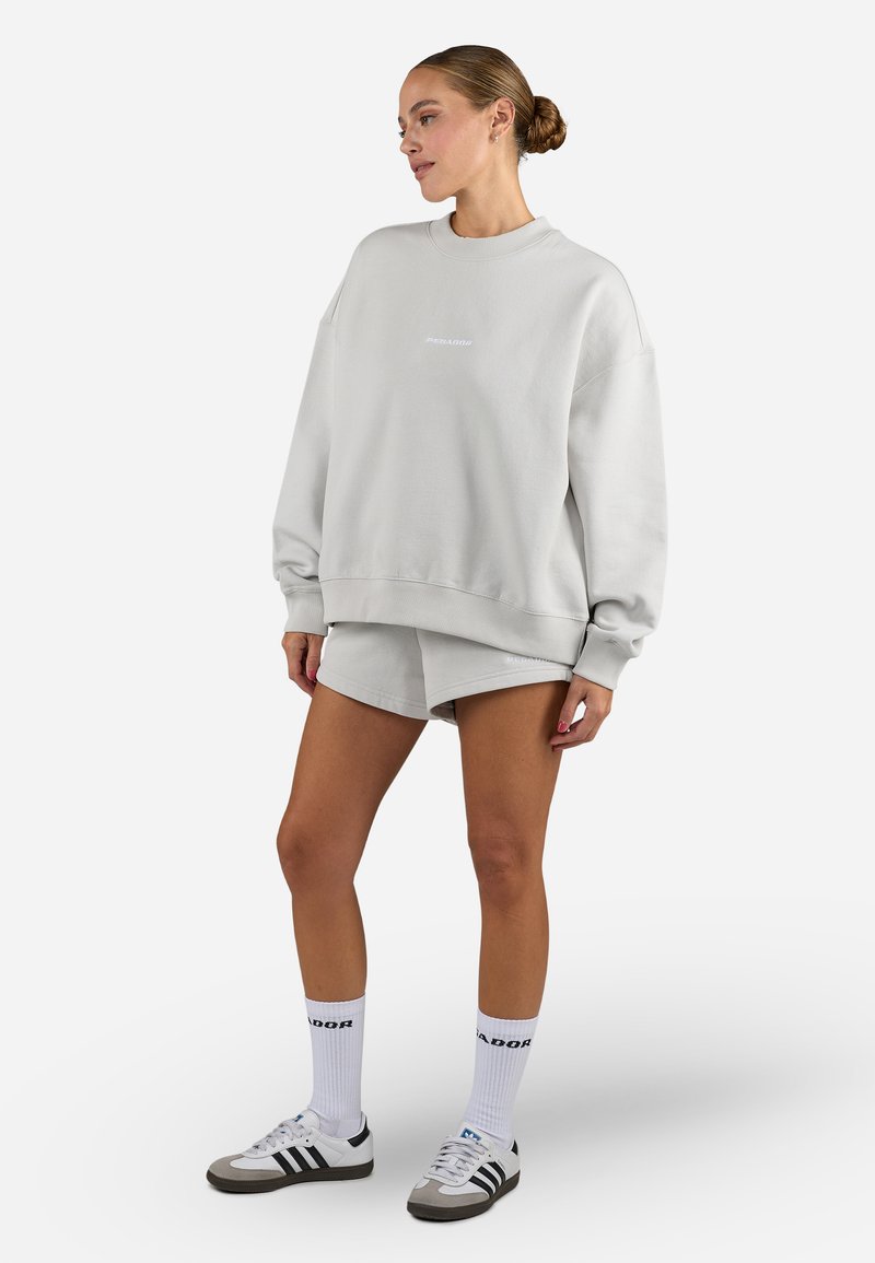 Light grey oversized sweatshirt with crew neck, and matching shorts. Features soft texture, ribbed cuffs, and subtle branding. Paired with white and black sneakers and socks.