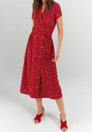 Femme portant une robe midi rouge à imprimé floral bleu, manches courtes, boutons sur le devant, et sandales rouges à talons hauts ouvertes à l'avant.