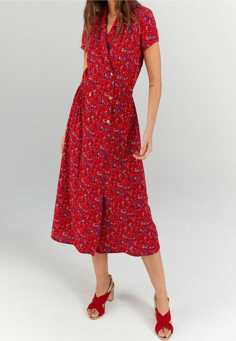 Femme portant une robe midi rouge à imprimé floral bleu, manches courtes, boutons sur le devant, et sandales rouges à talons hauts ouvertes à l'avant.