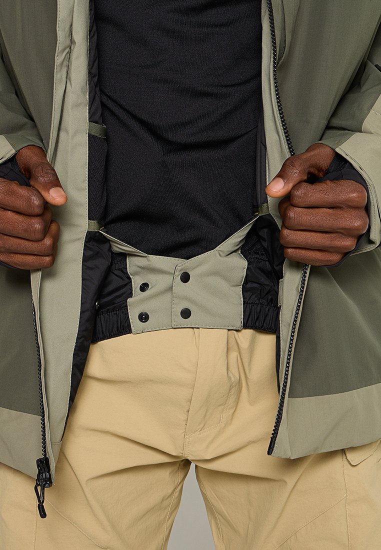 Chaqueta de cremallera verde y negra con un forro interior texturizado, cintura ajustable y puños elásticos, combinada con pantalones beiges.