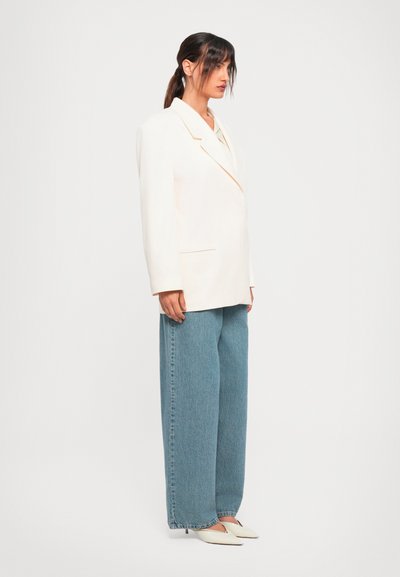 Vrouw die zijwaarts staat en een oversized witte blazer, wijde blauwe jeans en witte puntige hakken draagt, tegen een effen witte achtergrond.