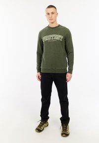 Olijfgroen sweatshirt met "PROTEST SPORTSWEAR" in crèmekleurige letters, gecombineerd met zwarte jeans en sneakers met een camouflagepatroon.