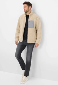 Giacca di pile beige con una tasca a zip grigia sul petto, maniche lunghe, camicia nera sotto, abbinata a jeans grigi e sneakers crema.