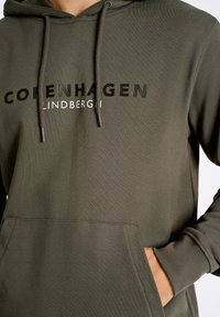 Felpa verde oliva scuro con tasca a marsupio frontale, cappuccio con coulisse e bold "COPENHAGEN LINDBERGH" in colori a contrasto.