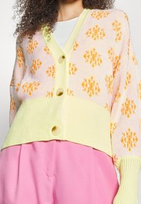 Vrouw draagt een gebreid vest in pastelgeel en roze met oranje bloemmotieven en grote knopen over een wit topje en roze broek.