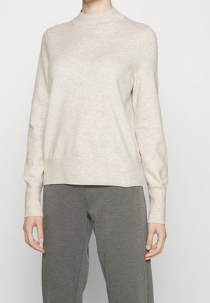 Žena oblečená v světle béžovém dlouhém pleteném svetru a volných šedých kalhotách, stojící před jednobarevným bílým pozadím.