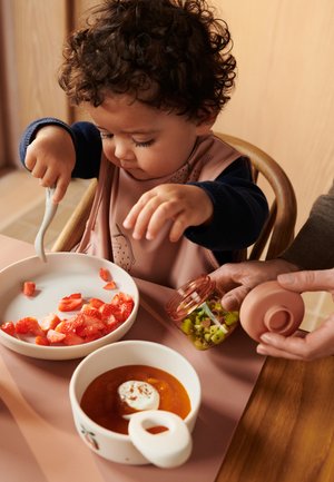 Un niño pequeño con un babero está sentado en una mesa comiendo fresas picadas con un tenedor mientras un adulto sostiene un frasco con kiwi picado.