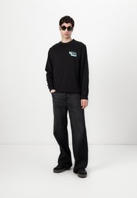 Zwarte crewneck sweatshirt met blauwe graphic op de borst, gecombineerd met donkergrijze wijdvallende jeans en zwarte sneakers. Model draagt zwarte zonnebril.