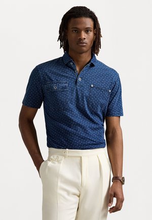 Korte mouwen marineblauwe polo met witte stippenpatroon, twee borstzakken en een knoopkraag. Gepaard met lichte geplooide broeken.