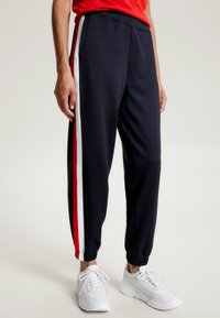 Marina joggerbyxor i ett mjukt tyg, med vertikala röda och vita sidostrepningar och elastiska muddar.Matchas med vita träningsskor.