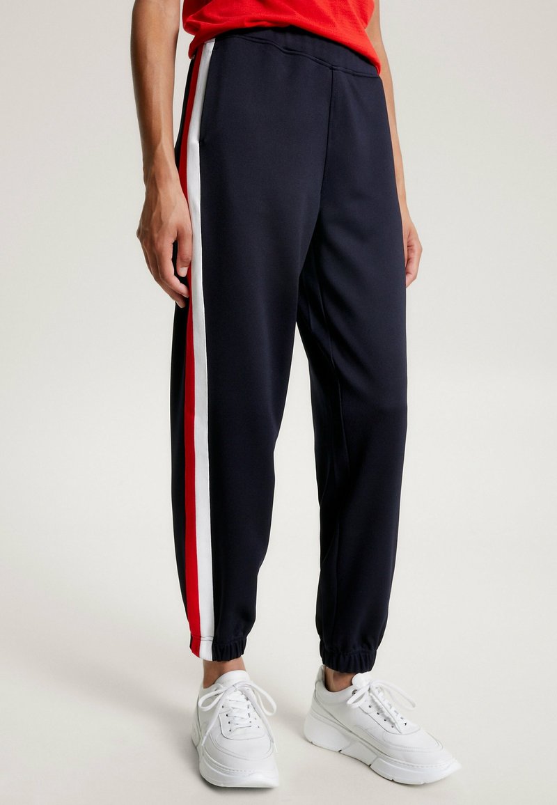 Marina joggerbyxor i ett mjukt tyg, med vertikala röda och vita sidostrepningar och elastiska muddar.Matchas med vita träningsskor.