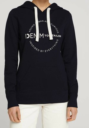 Personne portant un sweat à capuche noir avec des cordons blancs et un texte blanc circulaire indiquant "DENIM TOM TAILOR" et d'autres mots à l'avant.