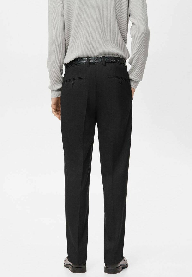 Person, die schwarze maßgeschneiderte Hose, schwarzen Gürtel, hellgraues, langärmliges, geripptes Hemd und schwarze Lederschuhe trägt, von hinten zu sehen.