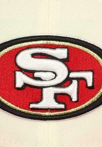 Cap features an embroidered red and white logo with black and gold outlines, showcasing the initials 'SF' in a bold design on a cream background.
