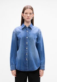 Camicia di denim in una tonalità blu medio, con chiusura frontale a bottoni, maniche lunghe e orlo curvo. Texture liscia con un design del colletto classico.
