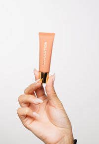 Peachfarget sminke-tube med gullbase, merket "REVOLUTION" med hvit tekst, holdt mellom fingrene med velstående negler.