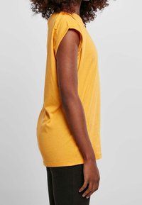 Nainen, jolla on kihara tukka, pukeutuneena keltaiseen lyhythihaiseen paitaan ja mustiin housuihin, seisoo sivuttain yksinkertaista vaaleaa taustaa vasten.