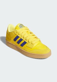 Basket jaune à tige basse avec des rayures bleues, de grosses lacets et une semelle beige, présentée sous un léger angle sur un fond clair.