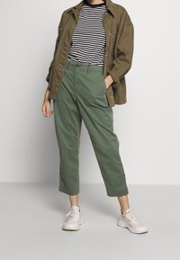 Olivově zelené, zkrácené kalhoty s volným střihem, kombinované s černobílým pruhovaným tričkem a zelenou pracovní bundou. Bílá sportovní obuv dotváří celý vzhled.