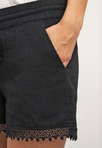 Czarne szorty z teksturowanej tkaniny, wyposażone w elastyczny pasek w talii, boczne kieszenie oraz koronkowe wykończenie z małymi pomponami.