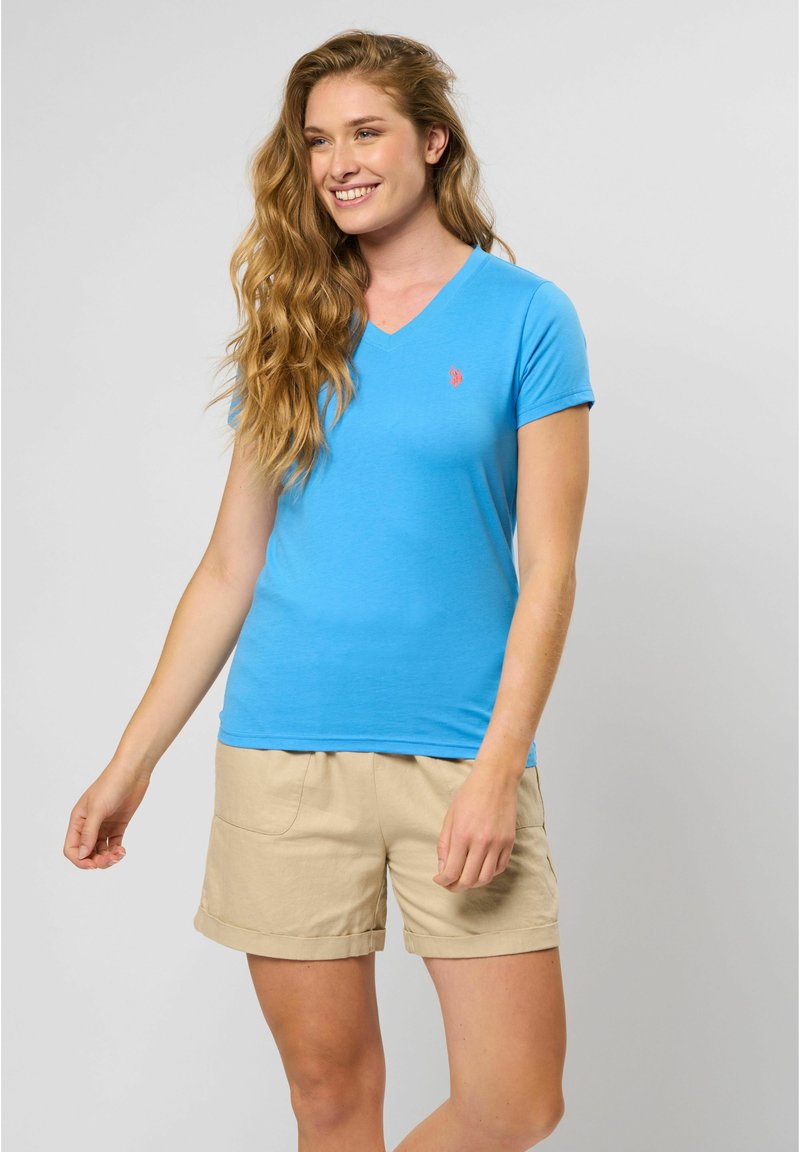 V-hals t-shirt met korte mouwen in felblauwe katoen, met een klein rood logo op de borst. G gedragen met kaki shorts.