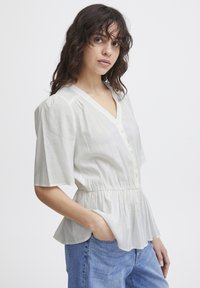 Blouse blanche légère avec un col en V et des manches courtes bouffantes. Présente un devant à boutons et une taille froncée, créant une forme peplum.