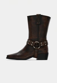 Καφέ σκούρα δερμάτινη μπότα μέχρι τον αστράγαλο με χαμηλό τακούνι, μυτερή μύτη και διακοσμητικό λουράκι με ασημένιες καρφίτσες και μεταλλικό κρίκο.