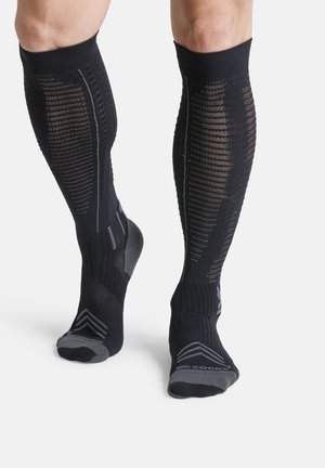 Benen met zwarte kniehoge compressiekousen met grijze versterkte tenen, hielen en ventilatiepatronen op kuiten en voeten.