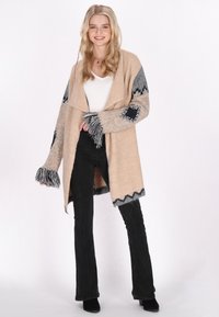 Beige oversized cardigan med grå accenter; har fransade ärmar och dekorativa pärlmönster; passar bra med svarta utsvängda byxor.