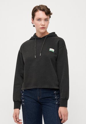 Svart kortermet hettegenser med snor, med et lite flerfarget merke på fronten. Kombinert med blå jeans med snøring på siden.