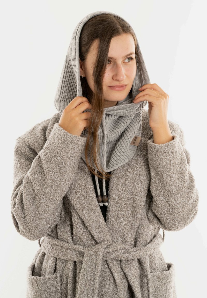 Vrouw past een lichtgrijze gebreide capuchon aan over haar hoofd, draagt een textuur beige mantelkostuum met riem en zakken, en kijkt iets opzij.