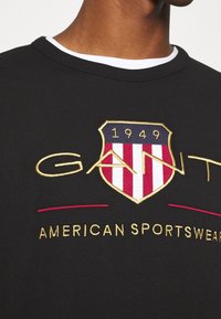 Sudadera negra con un escudo bordado en oro y rojo que incluye los textos "GANT" y "AMERICAN SPORTSWEAR", cuello redondo y tejido texturizado.
