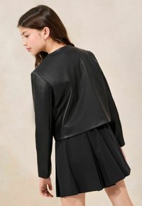 Schwarze, cropped Lederjacke mit langen Ärmeln, kombiniert mit einem gefältelten schwarzen Rock. Glatte Textur und minimalistisches Design ohne sichtbare Verzierungen.
