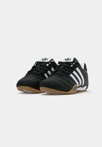 Adidași negri cu dungi albe, cu talpă din cauciuc și logo-uri pe limbă, prezentați dintr-un unghi joasă din față pe un fundal alb.