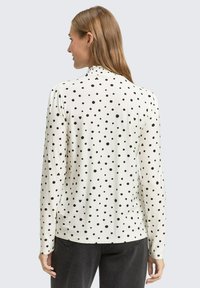 Weißes Langarmoberteil mit schwarzen Punkten. Verfügt über einen runden Ausschnitt und Raffung im Rückenbereich. Aus weichem, leichtem Material.
