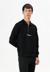 Crna zip-up dukserica s visokim ovratnikom i vezom "Original Studio" na prednjem dijelu. Mekana, teksturirana tkanina i opušteni kroj.