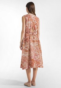 Vrouw gezien van achteren, draagt een mouwloze midi-jurk met oranje en roze bloemenprint, een open rug en een ceintuur in de taille, gecombineerd met beige schoenen.