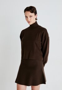 Brun strikket turtleneck-genser med senkede skuldre, kombinert med et matchende kort, utsvingt skjørt. Myk tekstur og minimalistisk design.