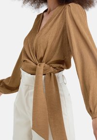 Top avvolgente marrone, con texture, maniche lunghe e un fiocco in vita, abbinato a pantaloni chiari a vita alta.