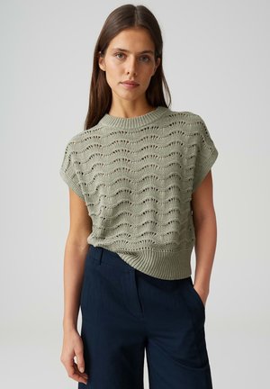 Jonge vrouw die een lichtgroen gebreid T-shirt met korte mouwen en een golfpatroon draagt, samen met een donkerblauwe broek met hoge taille, staand met één hand in de zak.