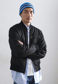 Schwarze Bomberjacke mit einer Reißverschlusstasche, über einem hellblauen Hemd und einem weißen Unterhemd getragen. Blau- und schwarzgestreifte Mütze. Lässige Pose.