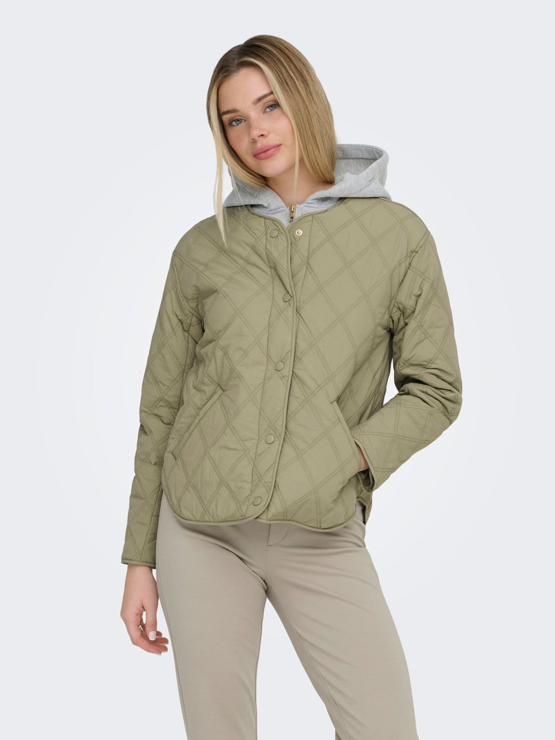 Ung kvinde med blondt hår iført en lysegrå hættetrøje under en quiltet olivengrøn jakke og beige bukser, stående med den ene hånd i lommen.