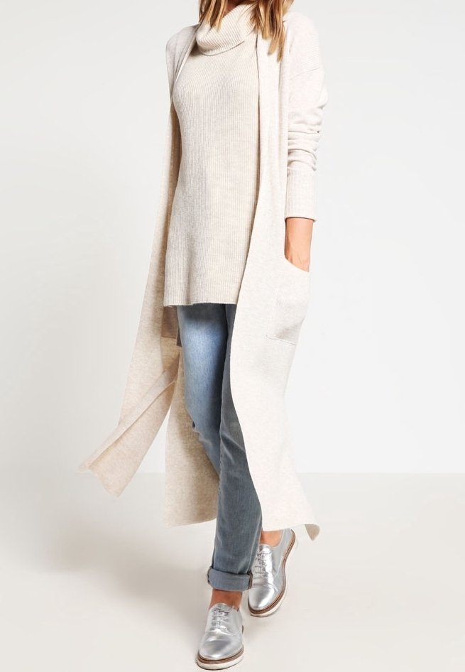 Femme portant un long cardigan beige, un pull à col roulé beige, un jean bleu retroussé et des chaussures argentées, marchant sur un fond uni.