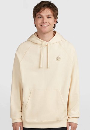 Jonge man met lichtbruin haar die een crèmekleurige hoodie draagt met voorzak en klein rond logo op de borst, glimlachend tegen een effen achtergrond.