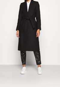 Abrigo negro largo con cinturón de lazo, cuello de solapa y cierre de botones, combinado con pantalones negros brillantes y zapatillas blancas.