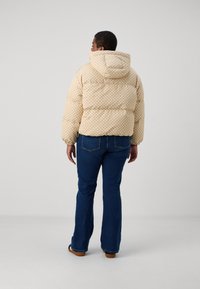 Giacca imbottita trapuntata di un beige chiaro con un motivo geometrico; presenta un cappuccio e polsini elastici, abbinata a jeans a zampa d'elefante blu.
