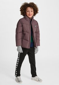 Brun vadderad jacka med hög krage, grå handskar, svarta byxor med vita chevronränder och silverfärgade sneakers. Barn med lockigt hår.