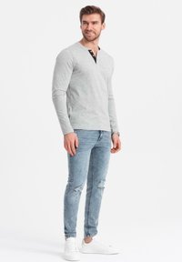 En man står klädd i en ljusgrå långärmad henley-tröja, slitna ljusblå jeans, vita sneakers och en armbandsklocka, leende och tittar åt höger.