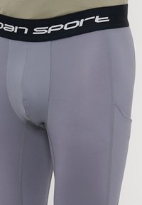 Šedé športové legíny s čiernym elastickým pásom, na ktorom je v bielej farbe nápis "sport". Materiál sa zdá byť elastický s hladkou textúrou.